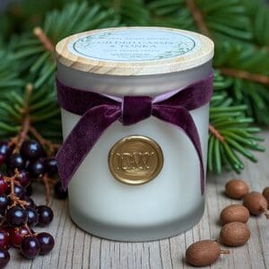 The Gilded Cassis & Tonka Candle, in a frosted glass jar with a gold wax seal and purple velvet ribbon, sits on wood amid dark berries, acorns, and pine branches. It features Tonka and Cassis notes and has a wooden lid with a decorative label.
