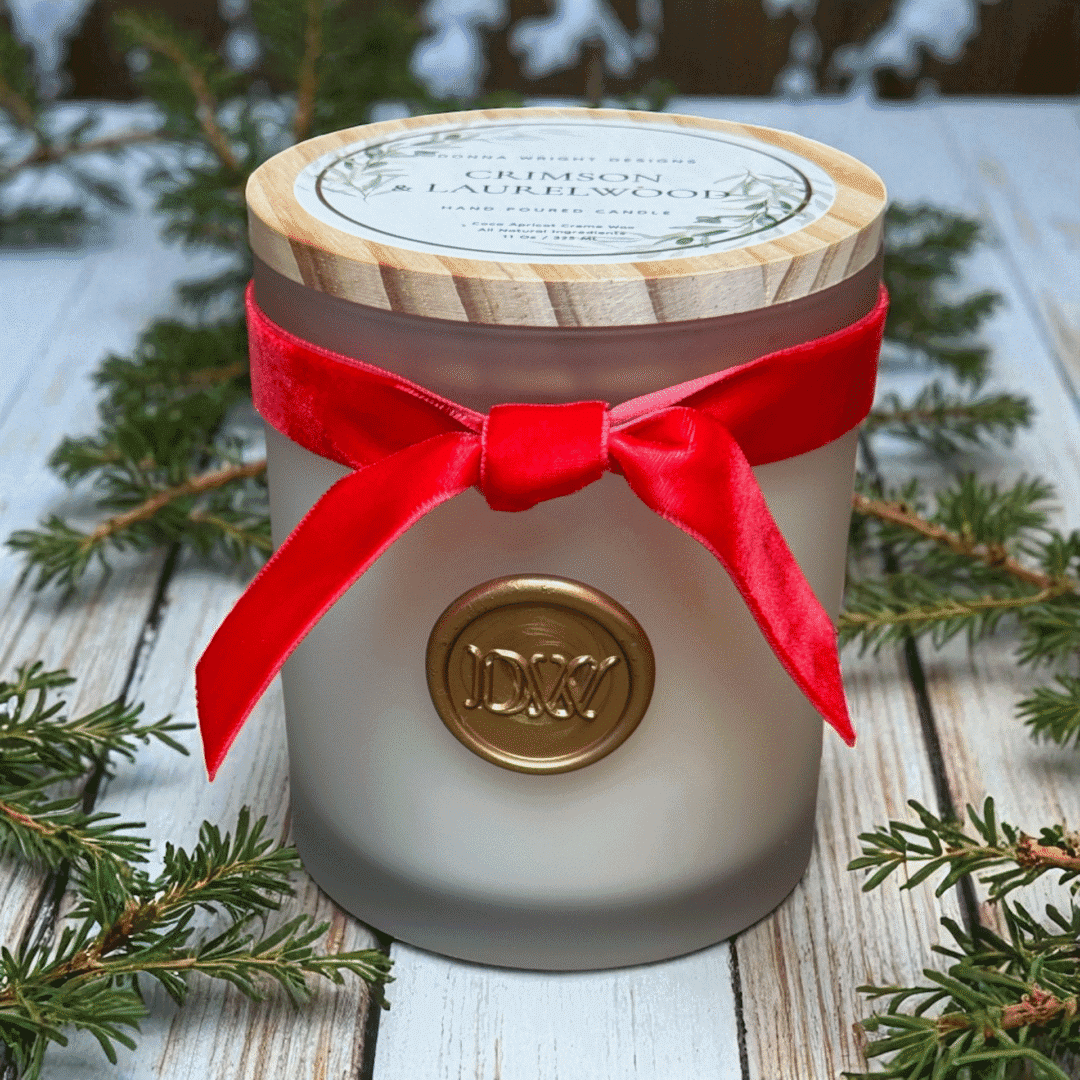A frosted glass jar candle from Crimson & Laurelwood Candles, tied with a red velvet ribbon and gold wax seal, rests on a wooden surface surrounded by pine branches. The label displays "Crimson & Laurelwood Candles.