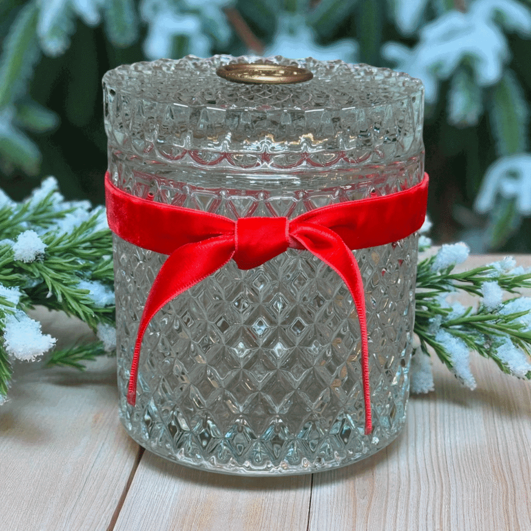A clear, textured glass jar with a lid, adorned with a red velvet bow, from Crimson & Laurelwood Candles, sits on a wooden surface surrounded by snowy pine branches.