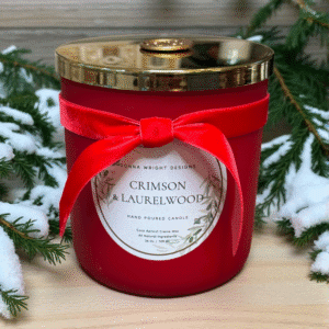 A red jar candle from Crimson & Laurelwood Candles, topped with a gold lid and red velvet bow, sits on a pine-adorned wooden surface—an inviting holiday display.