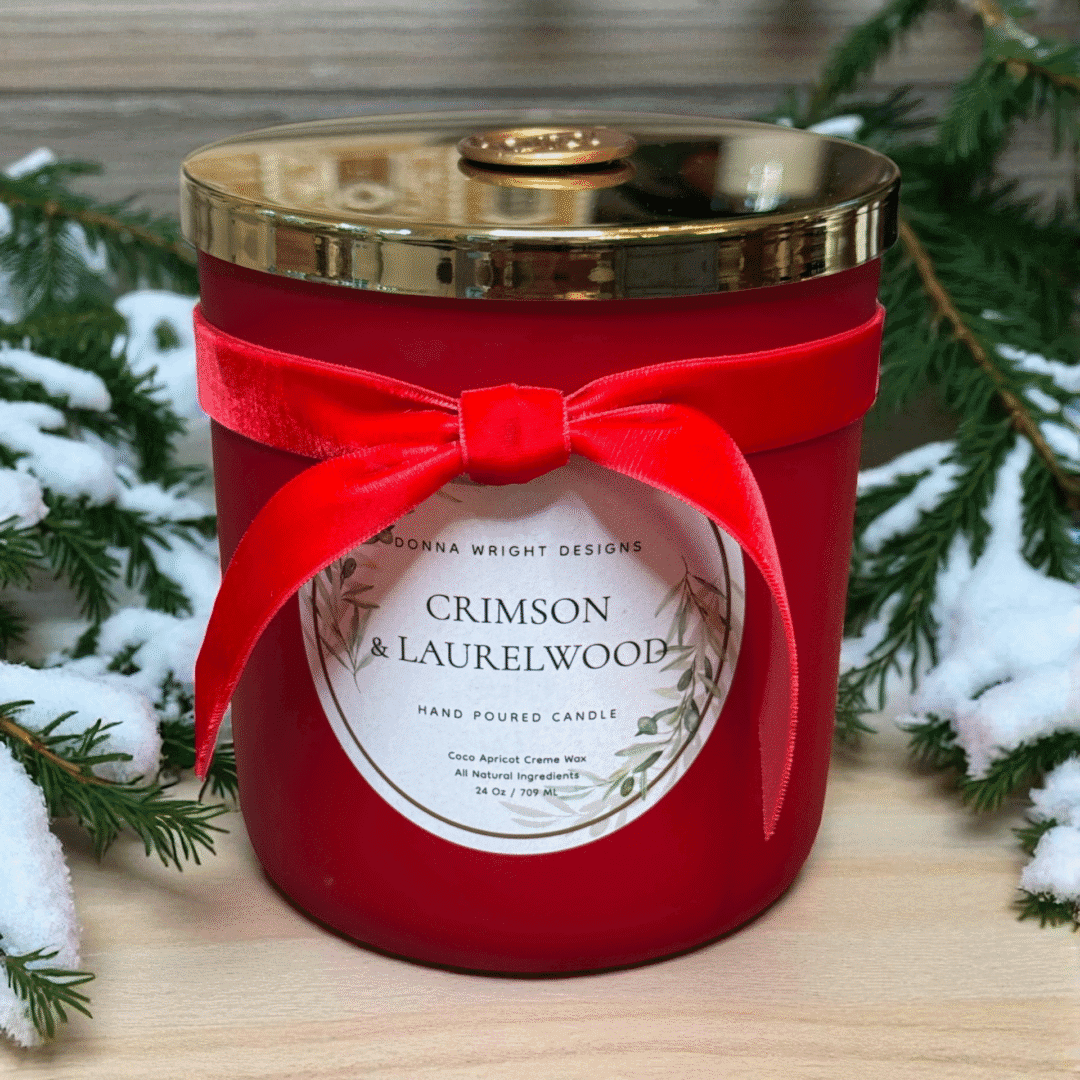 A red jar candle from Crimson & Laurelwood Candles, topped with a gold lid and red velvet bow, sits on a pine-adorned wooden surface—an inviting holiday display.