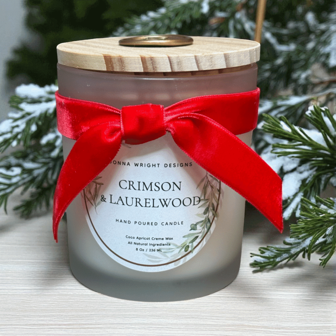 A Crimson & Laurelwood Candles frosted glass jar with wooden lid and red velvet bow sits before snow-dusted pine branches, evoking a festive holiday atmosphere.