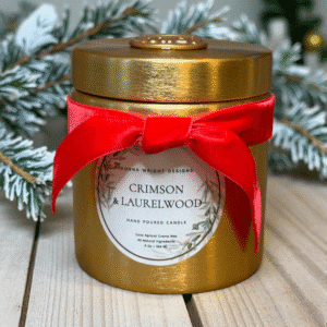 A gold candle jar with a red velvet bow rests on wood, labeled "Crimson & Laurelwood Candles." Pine branches and lush greenery adorn the background, making it a perfect addition to your collection.