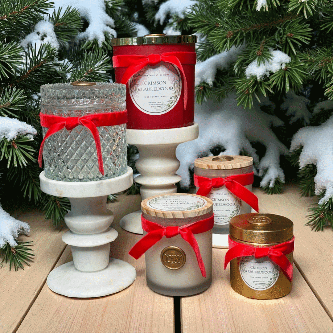 Five Crimson & Laurelwood Candles with red ribbons are arranged on white marble stands amid snow-dusted pine branches atop a wooden surface, evoking a festive holiday mood.