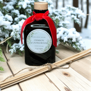 A scented diffuser labeled "Crimson & Laurelwood Candles" with a red velvet bow rests on wood beside reed sticks, capturing the cozy warmth and festive ambiance of the candle line among snow-dusted branches and trees.