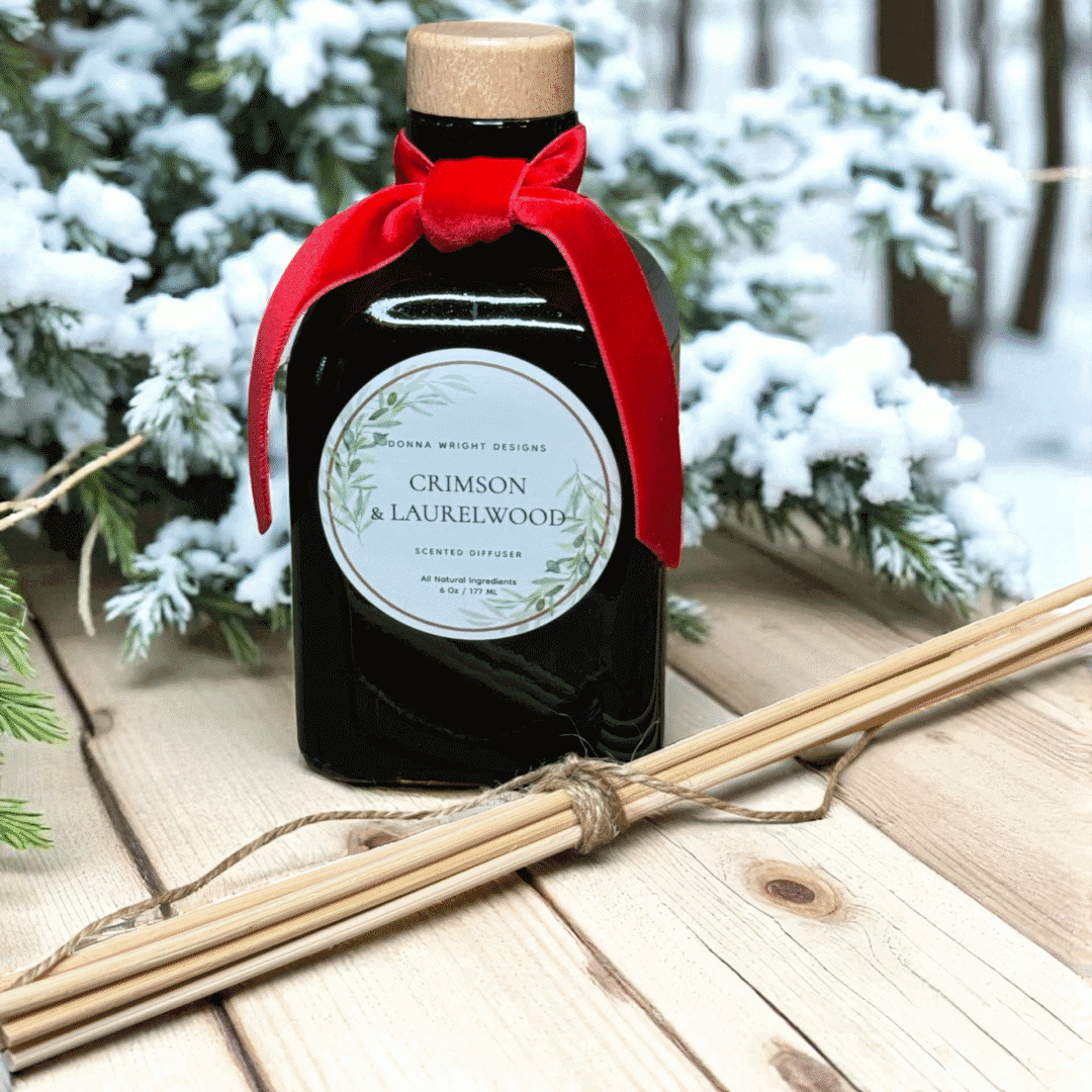 A scented diffuser labeled "Crimson & Laurelwood Candles" with a red velvet bow rests on wood beside reed sticks, capturing the cozy warmth and festive ambiance of the candle line among snow-dusted branches and trees.