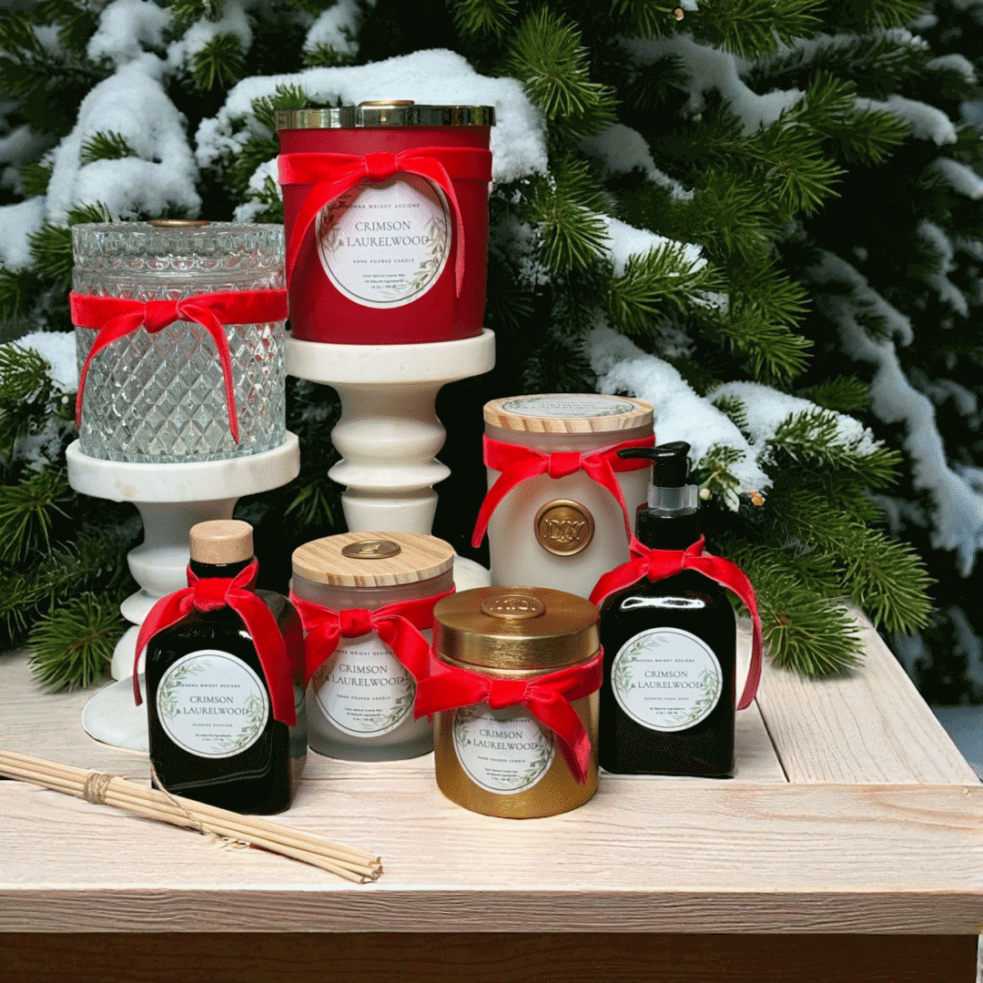 Crimson & Laurelwood Candles with red ribbons are displayed on a wooden table before snowy evergreens, creating a festive holiday scene.