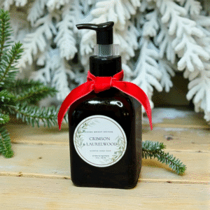 A dark pump bottle of scented hand soap from Crimson & Laurelwood Candles, adorned with a red ribbon and elegant botanical illustrations on the label, sits on a wooden surface in front of snowy pine branches.