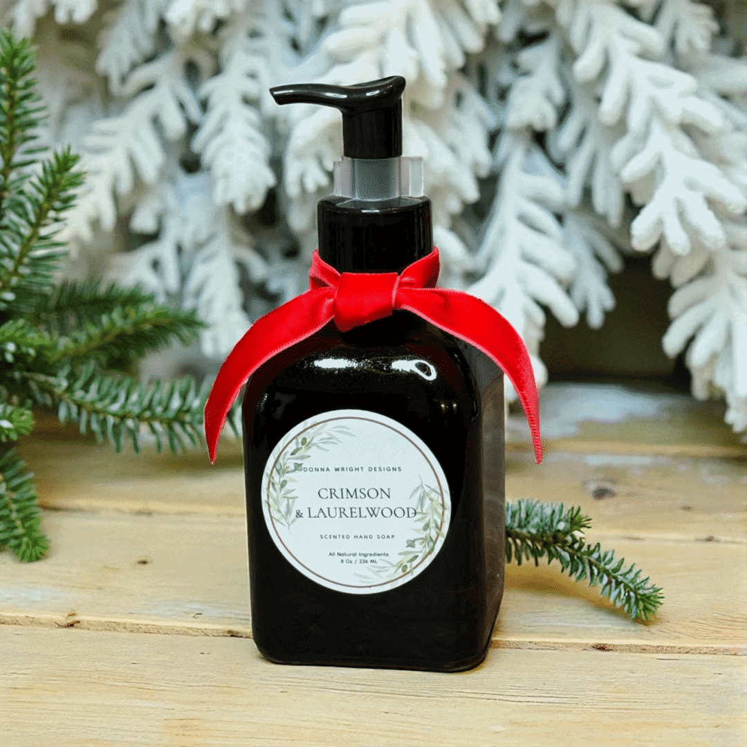 A dark pump bottle of scented hand soap from Crimson & Laurelwood Candles, adorned with a red ribbon and elegant botanical illustrations on the label, sits on a wooden surface in front of snowy pine branches.