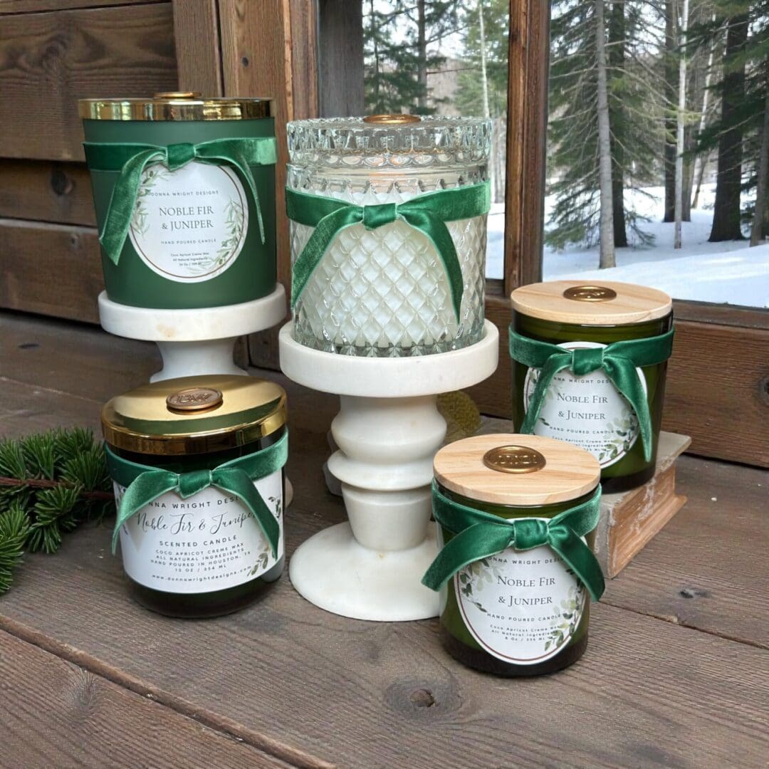 A display of five Noble Fir & Juniper candles with decorative labels and green ribbons by Donna Wright Designs, set on white stands and wooden surfaces, with a snowy forest visible through the window in the background.