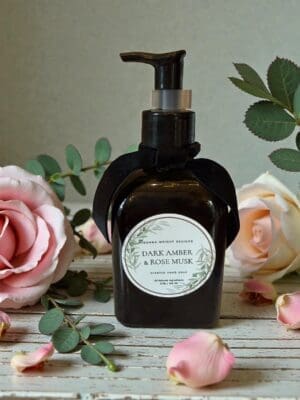 A Dark Amber & Rose Musk Hand Soap in a dark glass pump bottle sits on a rustic white wooden surface, surrounded by pink roses, eucalyptus leaves, and scattered rose petals.
