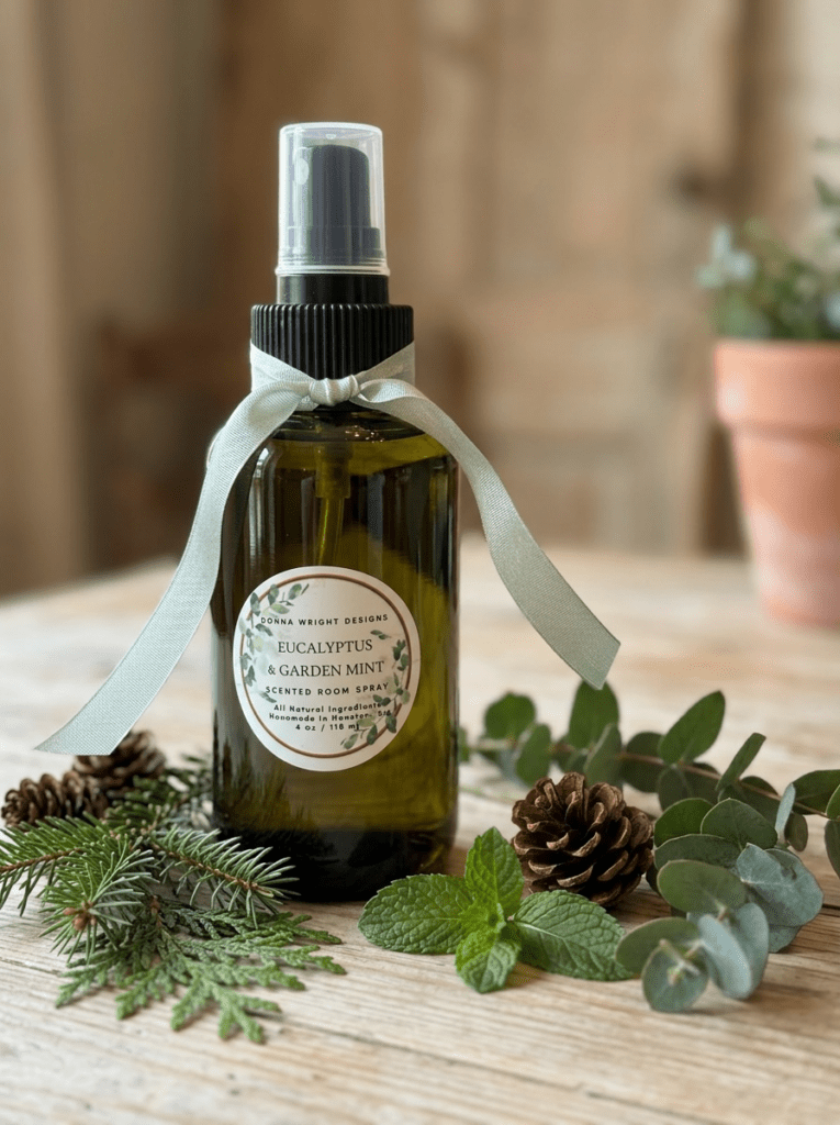 A green bottle of Eucalyptus & Garden Mint Room Spray by Donna Wright Designs, decorated with a light green bow, rests among pinecones, mint leaves, fir branches, and eucalyptus on wood—ideal for freshening any room.