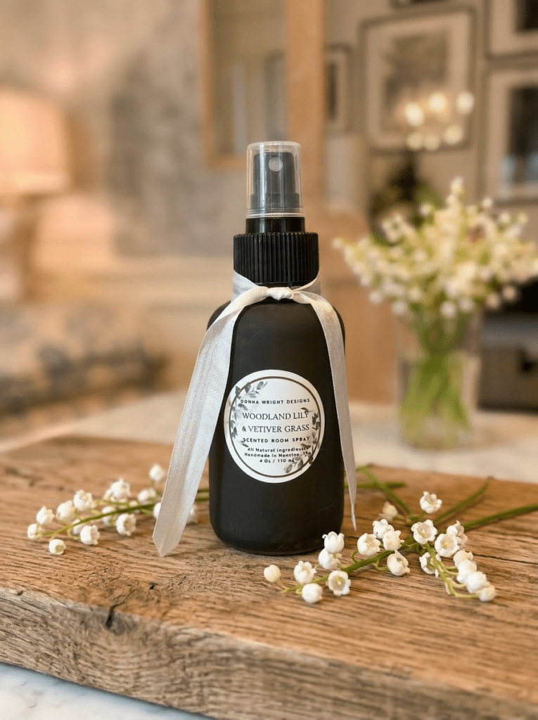 A dark spray bottle of Woodland Lily & Vetiver Grass Hand Soap with a white ribbon sits on rustic wood, surrounded by small white flowers. A cozy, blurred room appears in the background.
