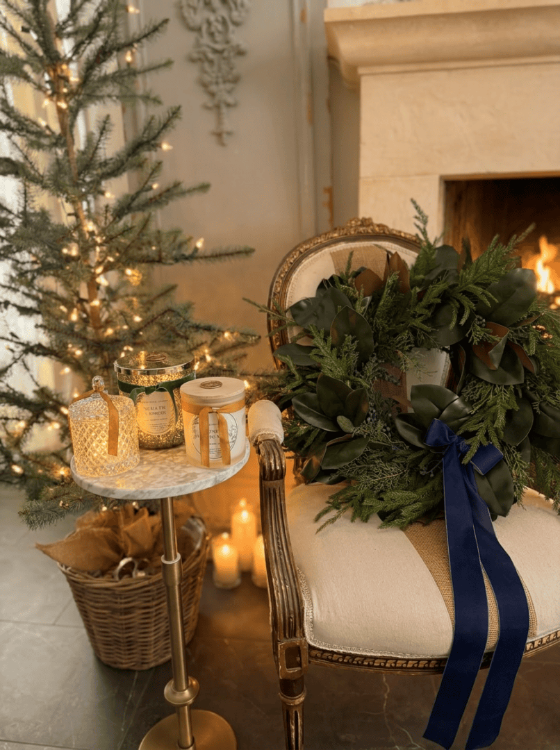 A cozy holiday scene with a lit fireplace, a decorated Christmas tree, and Donna Wright Designs candles with wooden wicks on a marble side table. A festive wreath with a dark blue ribbon graces an elegant armchair—explore Donna Wright Designs for more inspiration.