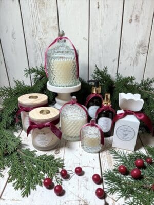 A holiday-themed display features Cranberry Yuletide Candles and luxury bath products in elegant glass jars and bottles, all adorned with red ribbons. The arrangement is set among pine branches and red berries on a white wooden background, with the rich seasonal scent of Cranberry Yuletide Candles enhancing the festive atmosphere.
