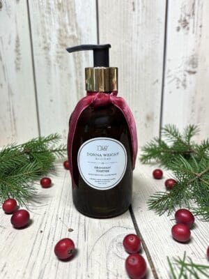 A bottle of Donna Wright Designs cranberry and vetiver hand wash, adorned with a red ribbon, rests on a white wooden surface surrounded by pine branches and cranberries, capturing the festive elegance of luxury holiday displays and evoking the ambiance of Cranberry Yuletide Candles.
