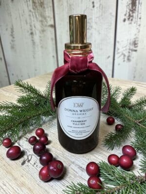 A dark glass spray bottle with a gold cap and red ribbon, labeled “Cranberry Yuletide,” is nestled among pine branches and cranberries on a wooden surface against a white rustic background—an elegant arrangement capturing the festive spirit of Cranberry Yuletide Candles.