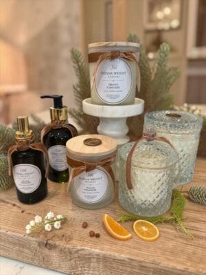 A collection of candles and skincare bottles, featuring the Orange Pomander Moisturizing Hand Wash, is arranged on a wooden surface adorned with greenery, dried orange slices, and decorative accents for a cozy indoor display.