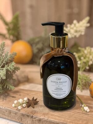 A green bottle of Orange Pomander Moisturizing Hand Wash by Donna Wright Designs, featuring a gold pump and brown ribbon, is displayed on a wooden surface surrounded by oranges, greenery, and delicate white flowers.