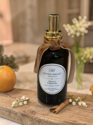 A bottle of Orange Pomander Moisturizing Hand Wash rests on a wooden surface with a satin bow, surrounded by oranges, a cinnamon stick, and small white flowers—making it the perfect choice for adding a harmonious touch of luxury to your home.