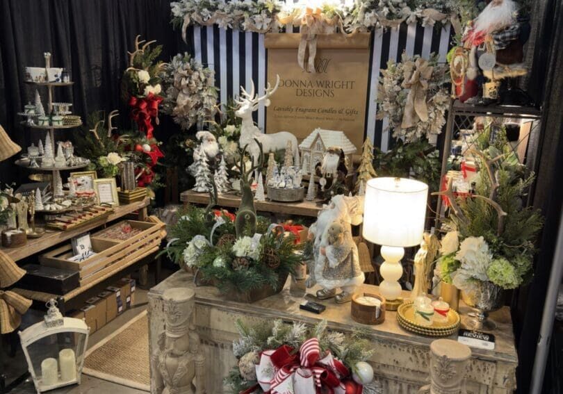A festive market booth displays Christmas decorations, including nutcrackers, wreaths, garlands, festive figurines, and holiday lights—perfect for those seeking elevated home fragrance and interiors. Greenery adorns the black-and-white striped backdrop and sign by Donna Wright Designs