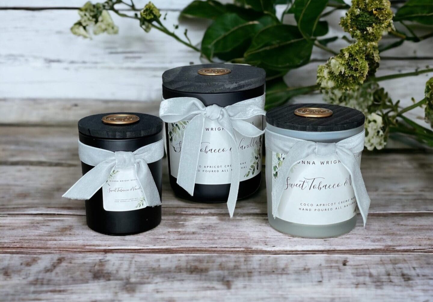 Three Donna Wright Designs Sweet Tobacco & Vanilla richly scented candles sitting on a table next to tobacco leaves.