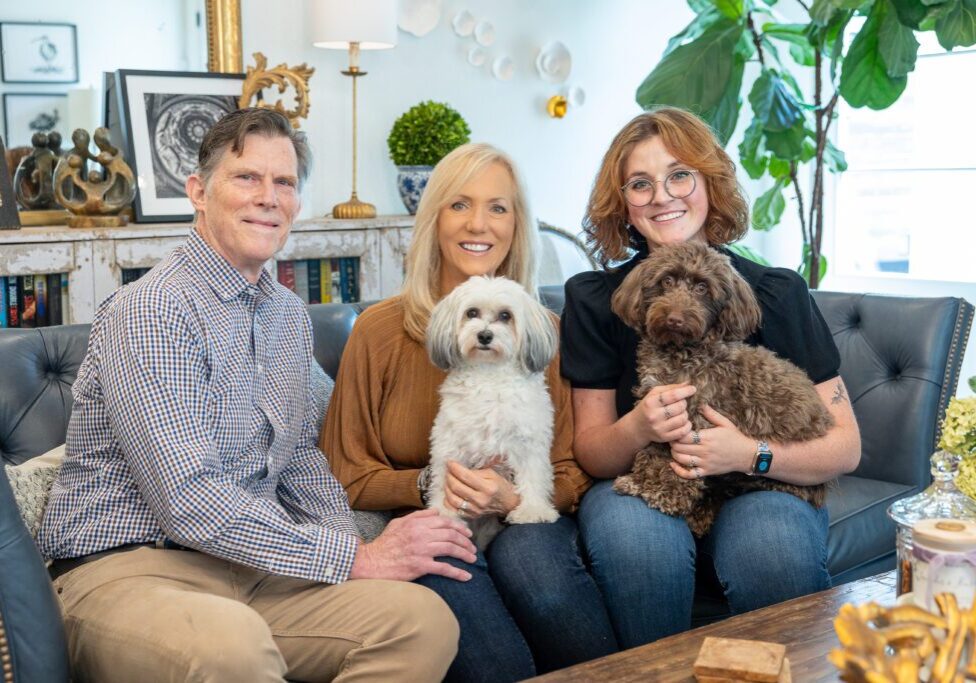 Donna Wright Designs Three smiling adults sit on a sofa in a cozy living room, holding two fluffy dogs, one man, two women, surrounded by elegant decor and houseplants, excitedly discussing the holiday markets 202