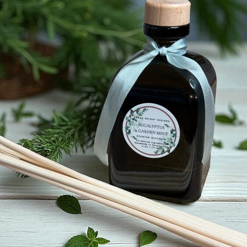 A dark bottle labeled “Eucalyptus & Garden Mint Hand Soap” with a wooden cap and blue ribbon rests on a white wood surface, surrounded by wooden reeds, mint leaves, and lush greenery in the background.