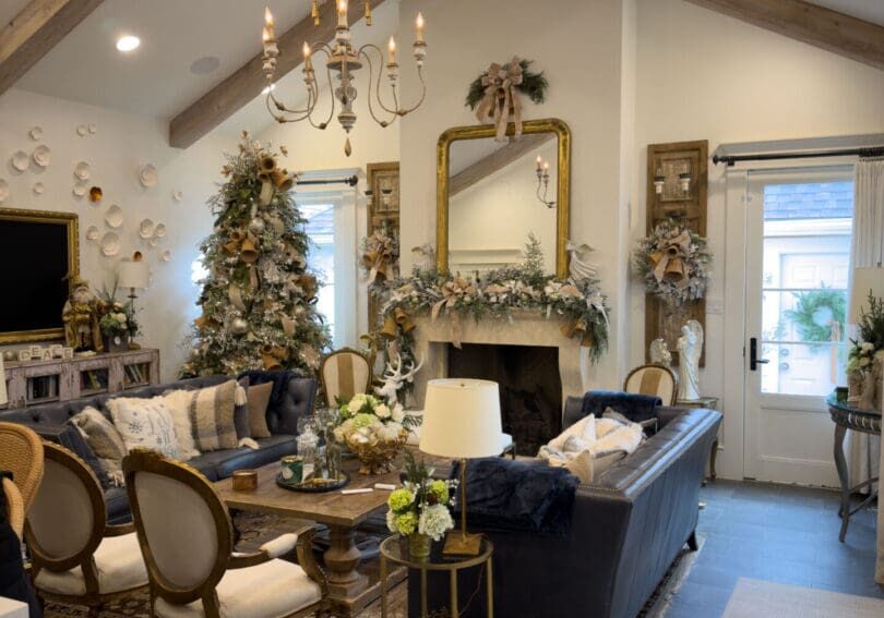 Elegant living room with vaulted ceiling, exposed wooden beams, a decorated Christmas tree, dark sofas, antique chairs, and floral arrangements. Donna Wright's touch is seen in the fireplace adorned with garlands and holiday décor for a warm, festive atmosphere.