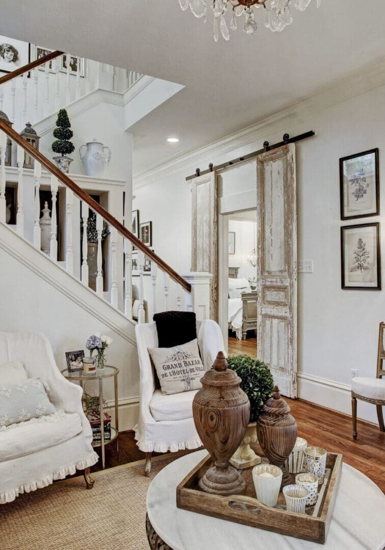 A cozy, elegant living room with vintage decor, white armchairs, a rustic coffee table, and a distressed blue dresser with mirror invites you in. The staircase is adorned with framed photos and greenery beneath a Copy of Header soft lighting glow.