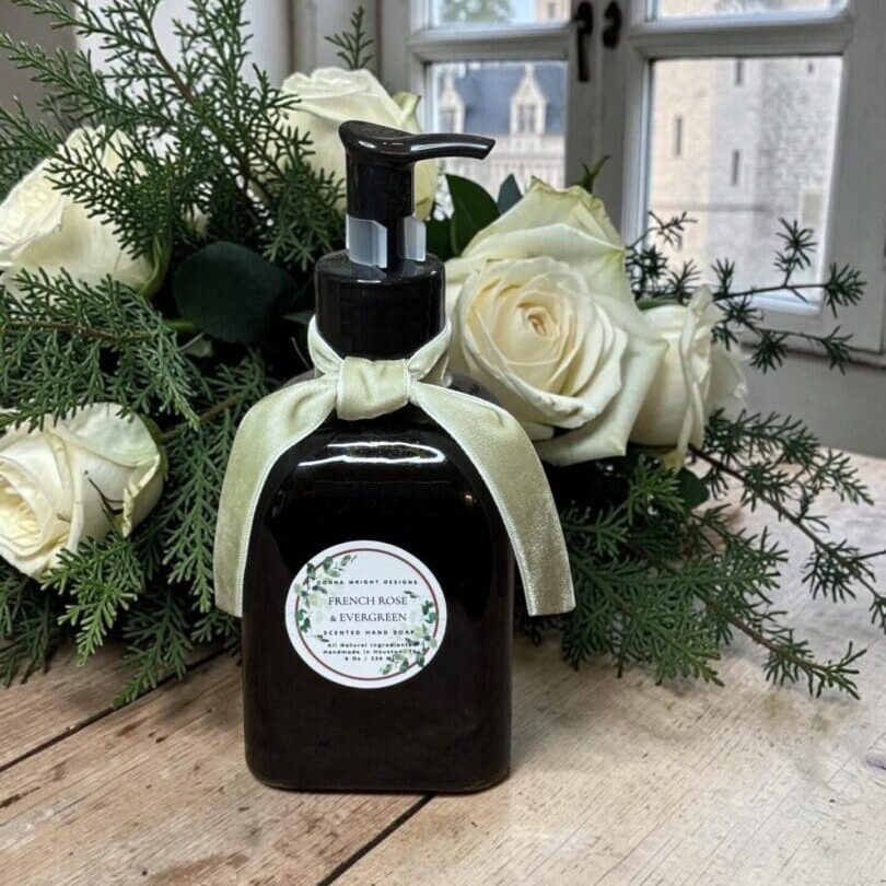 A dark pump bottle with a pale ribbon and floral label stands on a wooden surface near a window, the header of a stone castle visible outside, framed by white roses and greenery.