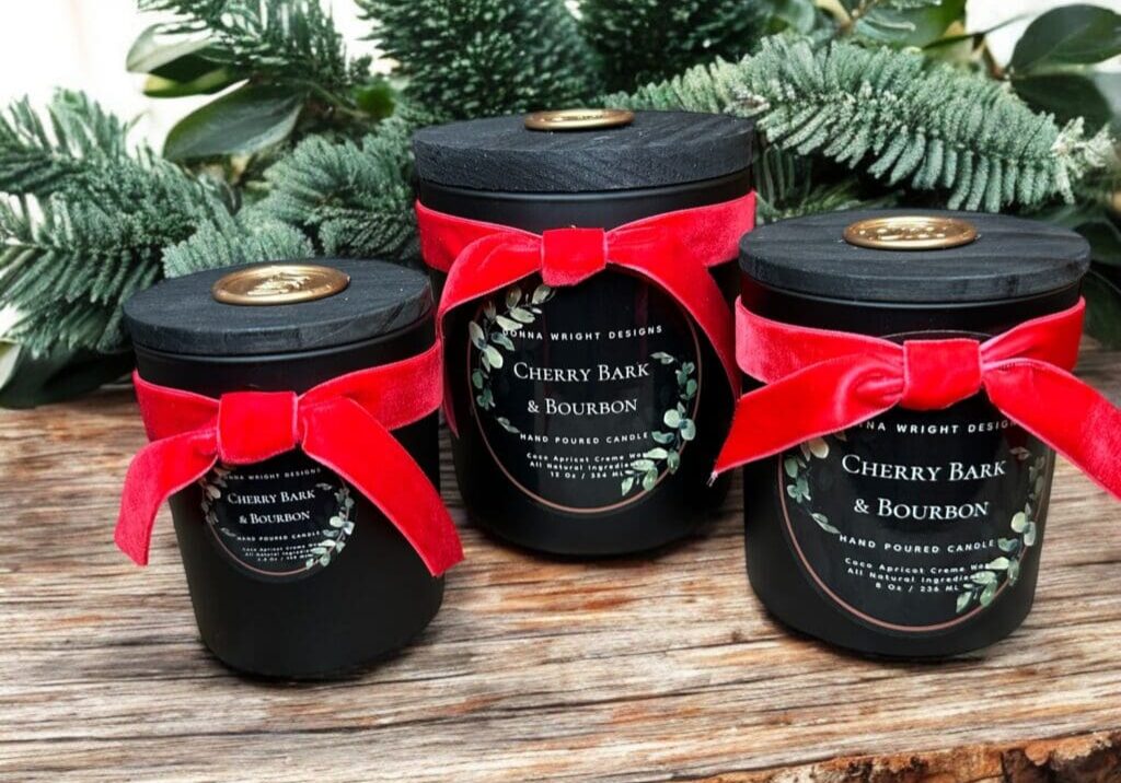 Three black candles labeled "Cherry Bark & Bourbon" are wrapped with red velvet ribbon and set on a rustic wooden surface adorned with evergreen branches. The candles vary in size, each with a gold seal on the matte black lid, creating a festive atmosphere reminiscent of the best fall and holiday markets.