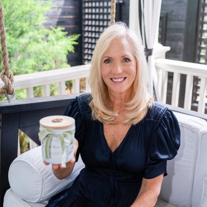 Donna Wright Designs A smiling, middle-aged woman with blonde hair, wearing a navy blue dress, sits on a swing chair on a porch holding a jar of our product toward the camera.