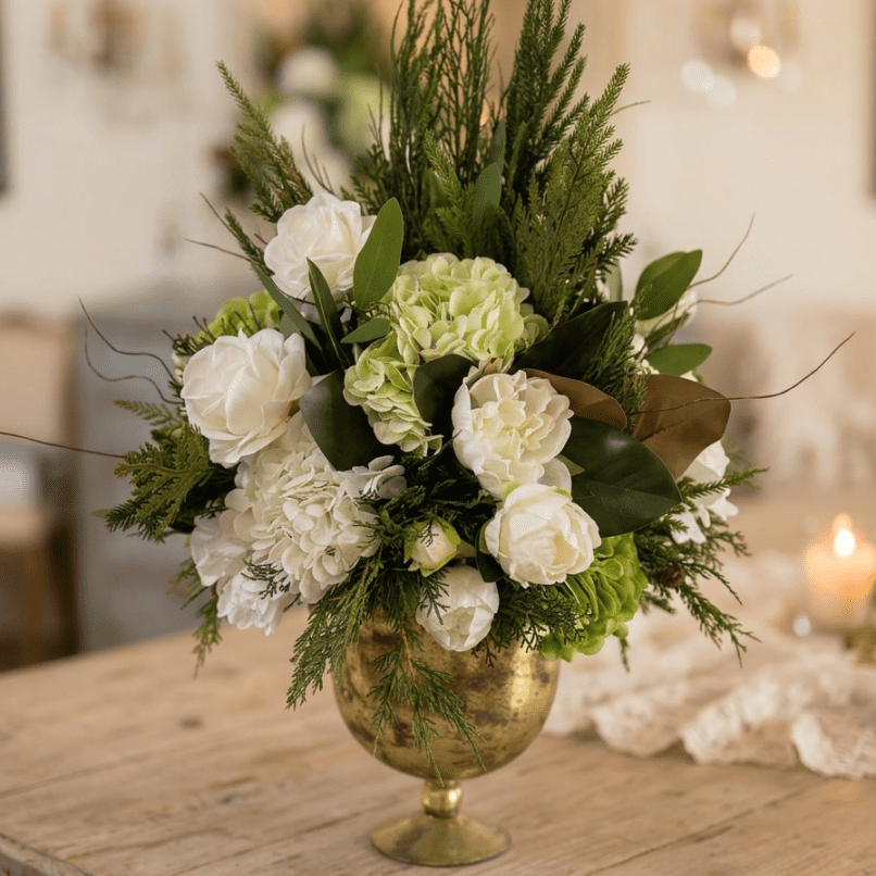 A gold vase filled with an arrangement of white and pale green flowers, assorted greenery, and leaves sits on a wooden table in a softly lit room with a blurred background.
