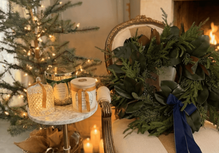 A cozy holiday scene with a lit fireplace, a decorated Christmas tree, and Donna Wright Designs candles with wooden wicks on a marble side table. A festive wreath with a dark blue ribbon graces an elegant armchair—explore Donna Wright Designs for more inspiration.
